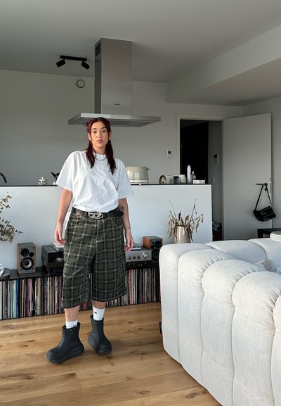 Camiseta blanca, corte extragrande, combinada con pantalones cortos de cuadro verde, botas de goma negras y calcetines blancos. Fondo interior con discos de vinilo y decoración.