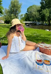 A person in a white floral tank top is seated on a white blanket, holding a pink cup. A wicker picnic basket and food containers are close by.