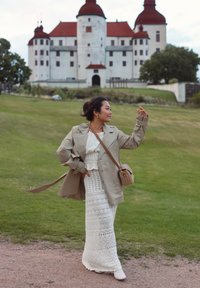 Light beige trench coat over a white lace maxi dress, accessorised with a small woven bag. Green grass and a castle with red roofs in the background.