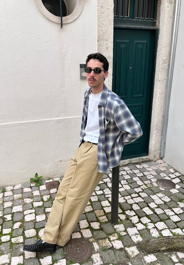 Junger Mann mit Sonnenbrille, karierter Hemd, weißem T-Shirt und beigen Hosen, der sich auf einen schwarzen Pfosten auf einer Kopfsteinpflasterstraße lehnt.