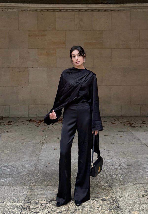 Woman standing against a beige tiled wall wearing all-black satin outfit, holding a black handbag with gold clasp in her right hand.