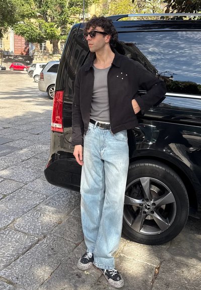 Chaqueta negra corta con botones visibles, camisa gris ajustada, jeans azul claro holgados y zapatillas negras con cordones blancos, de pie junto a un vehículo negro.