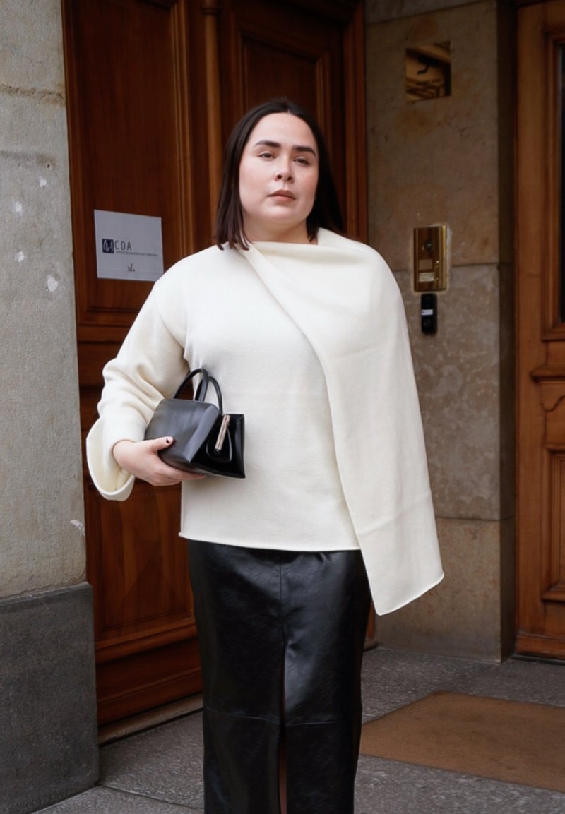 Frau steht vor einer Holztür, trägt ein weißes asymmetrisches Oberteil, einen schwarzen Lederrock und hält eine schwarze Handtasche.
