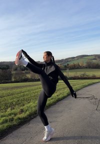 Fekete atlétikus trikó, fekete leggings és kesztyűk. Egy lábon áll, miközben a másikat felemeli. Futócipők rózsaszín és fehér díszítéssel. Zöld mezők a háttérben.