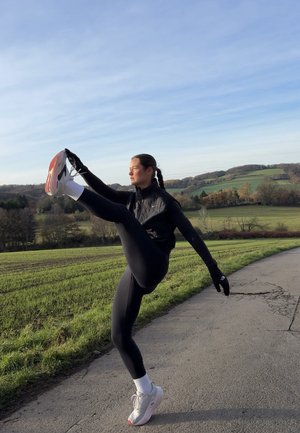 Chaleco deportivo negro, leggings negros y guantes. De pie sobre una pierna, levantando la otra. Zapatillas deportivas con detalles en rosa y blanco. Campos verdes al fondo.