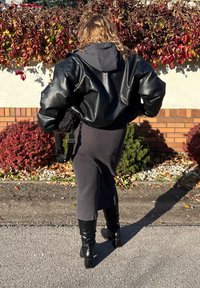 Giacca oversize in pelle nera con cappuccio, indossata sopra un vestito grigio a costine, abbinata a stivali neri al ginocchio con tacco, sullo sfondo colorato.