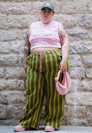 Top corto de crochet rosa, pantalones de pierna ancha a rayas verdes, bolso rosa y zapatos de plataforma. Tatuajes notables visibles en los brazos. Fondo de pared de piedra.