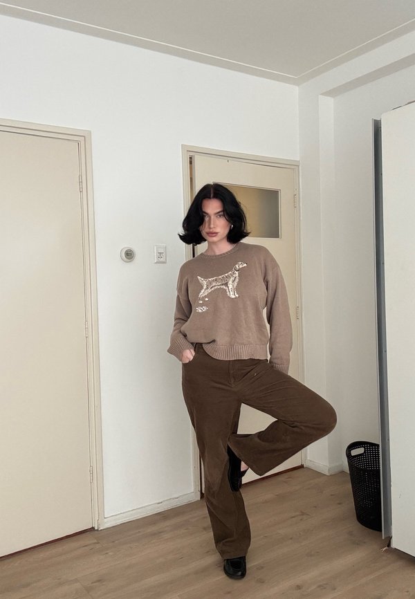 Vrouw staat op één been, draagt een beige trui met een hondmotief, bruine broek en zwarte schoenen, in een minimalistische kamer met lichte muren en houten vloer.