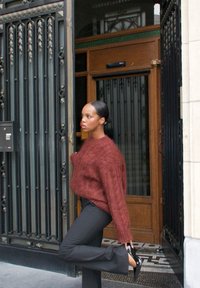 Maglione lavorato a maglia color ruggine con una vestibilità ampia, abbinato a pantaloni neri attillati. La persona indossa scarpe col tacco nere ed è in piedi vicino a una porta.