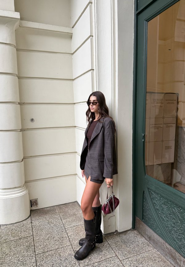 Woman wearing dark sunglasses, gray blazer, brown top, short skirt, and black boots, standing against a white column near a green door.