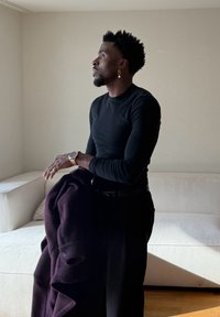 Un homme portant une chemise noire à manches longues tient un manteau sombre, debout près d'un canapé blanc dans une pièce minimaliste éclairée naturellement.