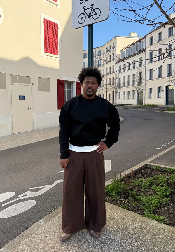 Homme avec coiffure afro portant un pull noir et un pantalon large marron, debout dans la rue près d'une piste cyclable et d'un bâtiment aux volets rouges sous un ciel dégagé.