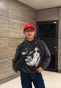 Felpa con cappuccio nera e grigia con stampa grafica, jeans blu, borsa a tracolla nera, cappellino rosso e occhiali da sole con lenti marroni su sfondo testurizzato.