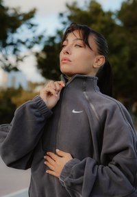 Fiatal nő a szabadban, a szürke Nike polárdzsekije cipzárját igazítja, a háttérben elmosódott fák láthatók.