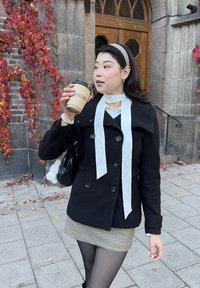 Jonge vrouw in een zwarte jas en een hondenstoethrok, die een koffiekop vasthoudt en staat op een stenen trottoir met herfstbladeren en rode wijnstokken.