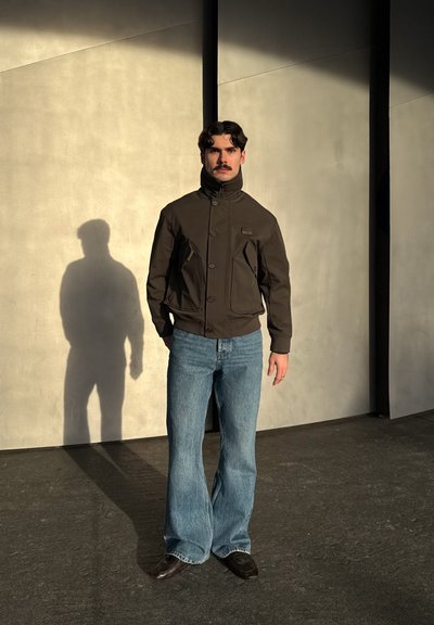 Hombre con bigote que lleva chaqueta marrón oscuro y vaqueros azules, de pie contra una pared beige con una sombra larga bajo la luz del sol.