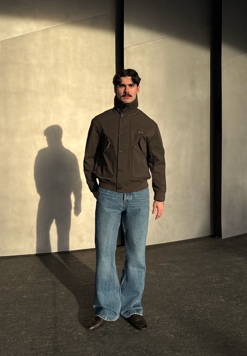 Homme moustachu portant une veste marron foncé et un jean bleu, debout contre un mur beige avec une longue ombre au soleil.