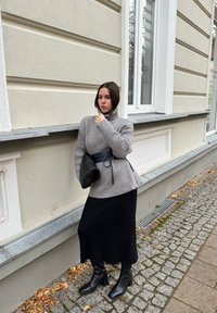 Woman in a grey turtleneck sweater, black midi skirt, and black boots stands on a cobblestone street next to a beige building, holding a black clutch.