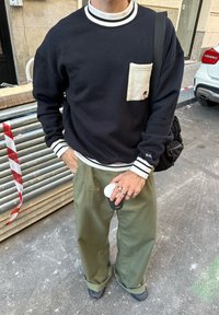 Persona con sudadera negra con bolsillo y franjas blancas, pantalones verdes, sosteniendo una taza de café cerca de una calle con un coche aparcado y barreras de metal.