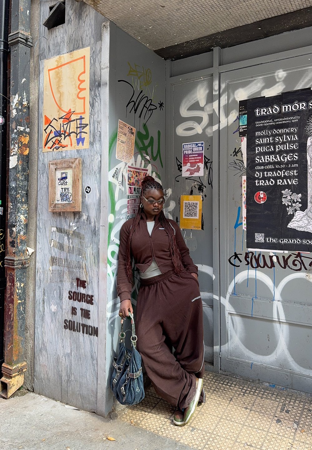 A person in a brown tracksuit and sunglasses leans against a graffiti-covered wall adorned with posters, holding a large handbag on a street corner in the city.