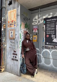 A person in a brown tracksuit and sunglasses leans against a graffiti-covered wall adorned with posters, holding a large handbag on a street corner in the city.