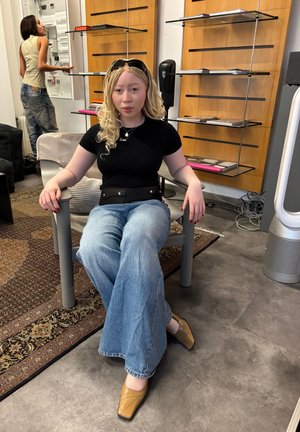 Una persona está sentada en una silla gris, vistiendo una camiseta negra y jeans acampanados de color azul claro. Lleva zapatos amarillos llamativos y un cinturón negro con detalles plateados.