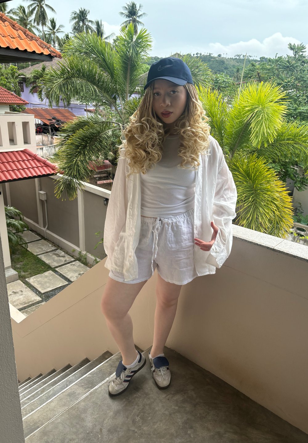 Blazer de linho branco sobre uma blusa branca e calções, com um boné azul-marinho e ténis cinzentos. O fundo apresenta vegetação e degraus de pedra.