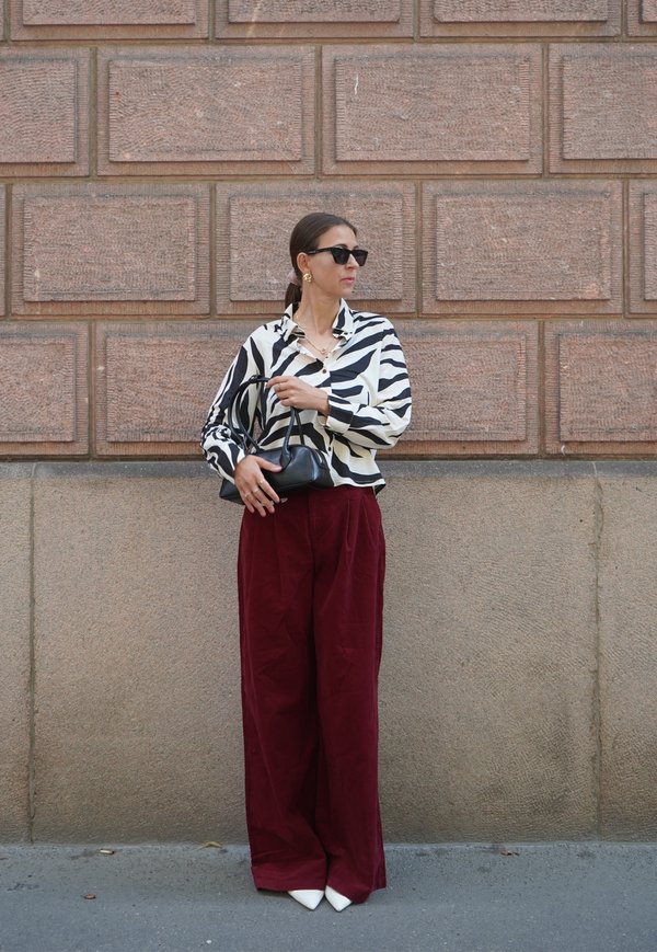 Woman wearing black and white patterned shirt, maroon wide pants, white shoes, and black sunglasses, holding a black handbag by a brick wall.