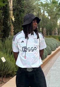 Λευκή ποδοσφαιρική φανέλα Ajax με μαύρες ρίγες και λογότυπο "Ziggo", συνδυασμένη με μαύρο καπέλο bucket και σκούρο τζιν. Υπαίθρια τοποθεσία με πράσινο τοπίο.