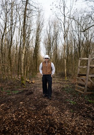 Persona con sombrero blanco, gafas de sol, chaleco marrón y pantalones negros caminando por un sendero de tierra a través de un bosque sin hojas en invierno.