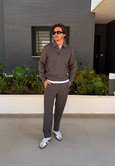 Suéter de punto gris con cuello y pantalones grises a juego. Combinado con zapatillas blancas y grises. El escenario presenta plantas verdes y una pared gris.