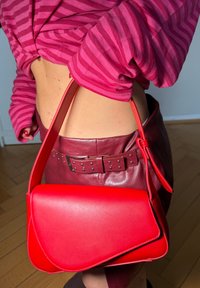 Sac à main en cuir rouge porté à l'épaule avec un haut rose à rayures et une jupe en cuir bordeaux avec ceinture cloutée sur un sol en bois.