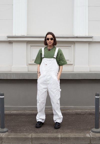 Un overol blanco de mezclilla, con tirantes ajustables y bolsillos frontales, combinado con una camiseta verde y zapatos negros.