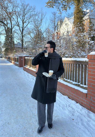 Hombre con abrigo negro, bufanda y guantes de pie en una acera nevada bebiendo café, con casas y árboles sin hojas al fondo en un claro día de invierno.