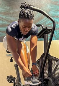 Svart treningssykkel med vifte, svart og sølv ramme, en person som knytter skoene. Bakgrunnen har dempede farger og teksturer.