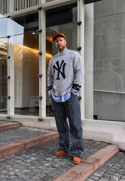 Hombre con gorra naranja, suéter gris con logo azul marino, pantalones azules a rayas y zapatos naranjas, de pie sobre escalones de adoquines frente a un edificio moderno de vidrio.