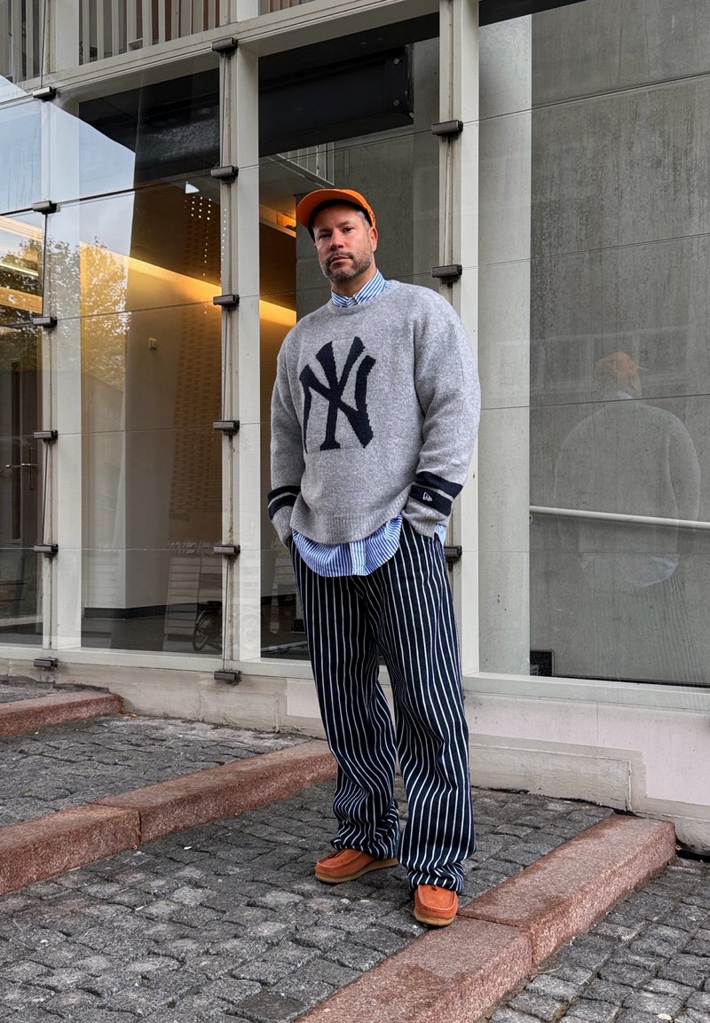 Muž v oranžovej čiapke, sivom svetri s námorníckym logom, modrých pruhovaných nohaviciach a oranžových topánkach stojí na kamenných schodoch pred modernou sklenenou budovou.