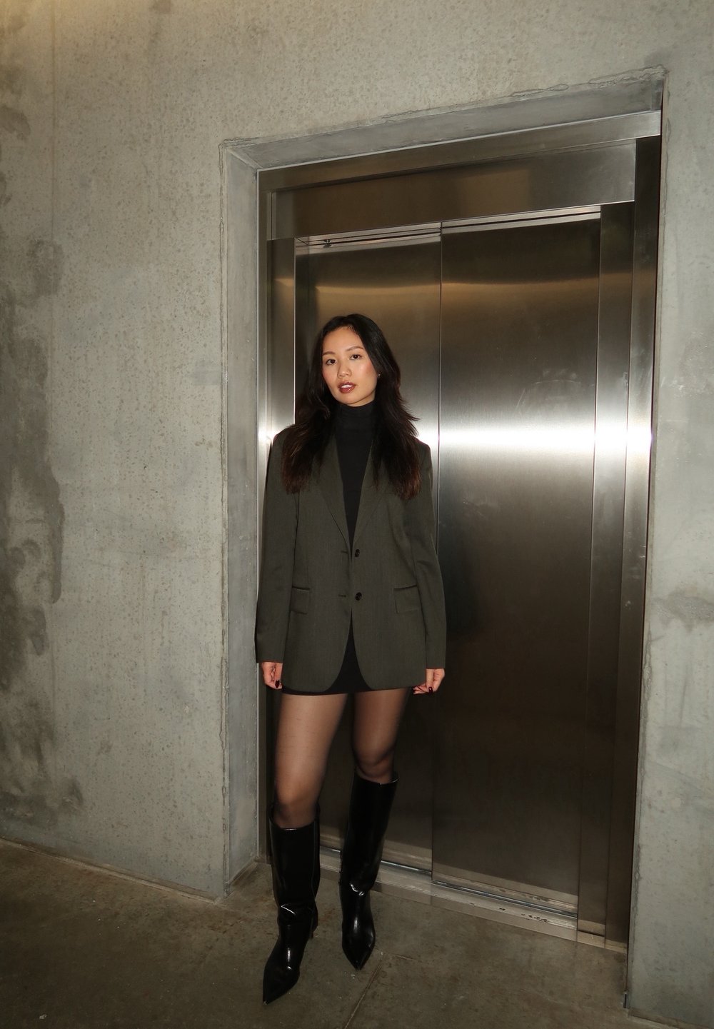 Õlivaheline bleiser musta seotud kaelusega, koos musta mini seeliku ja läikivate põlvini ulatuvate saapatega, betoonseina ja lifti taustal.