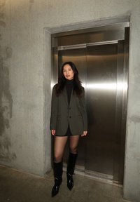 Õlivaheline bleiser musta seotud kaelusega, koos musta mini seeliku ja läikivate põlvini ulatuvate saapatega, betoonseina ja lifti taustal.