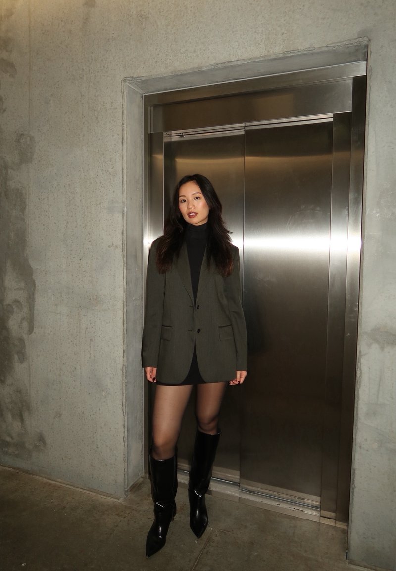 Olivno zelena blejzer preko črnega puloverja z visokim ovratnikom, v kombinaciji s črno mini krilo in sijajnimi škornji do kolen, pred betonsko steno in dvigalom.