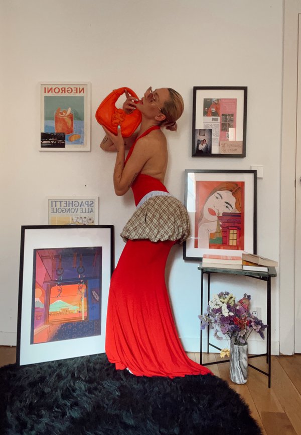 Personne en robe rouge posant en tenant un sac à main orange près d'une œuvre encadrée et d'une petite table avec des livres et des fleurs dans un espace de vie.