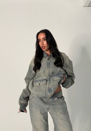 Mujer con el cabello largo y oscuro que lleva una chaqueta vaquera desgastada y grande y unos vaqueros a juego, sobre un fondo liso y claro.