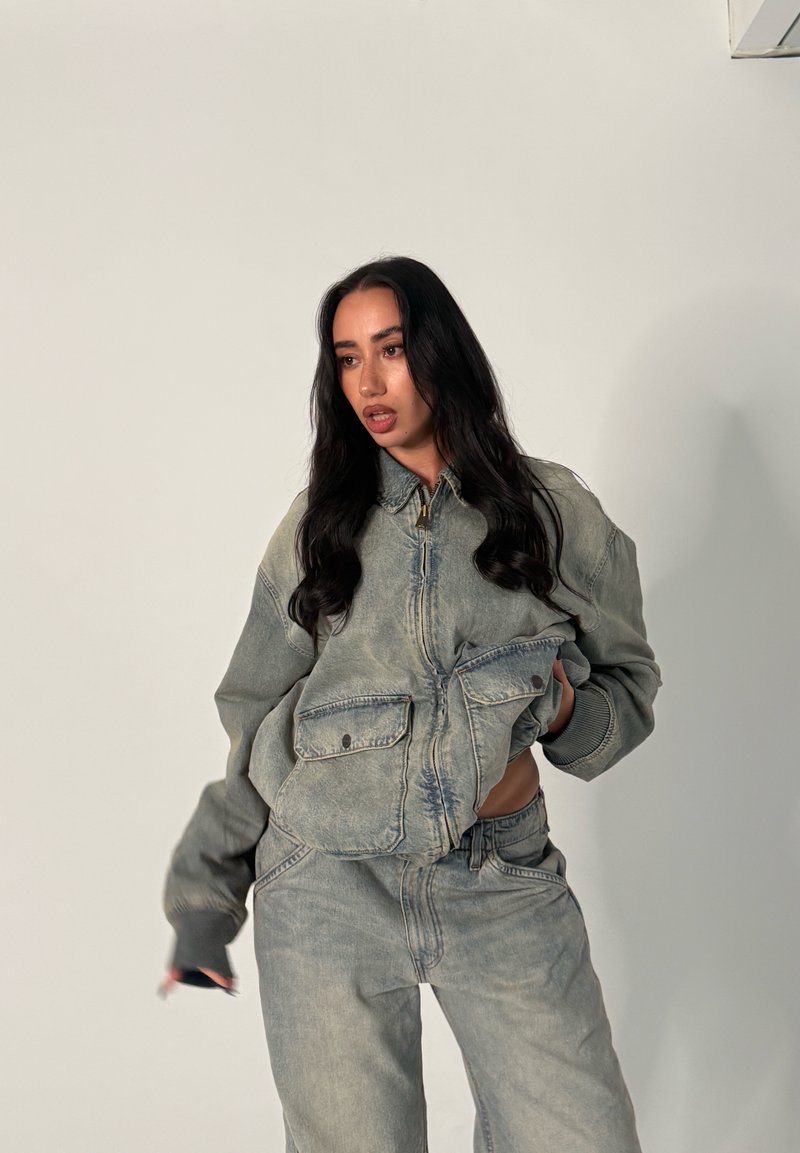 Kvinde med langt mørkt hår iført en stort oversized falmet denimjakke og matchende jeans mod en ensartet lys baggrund.