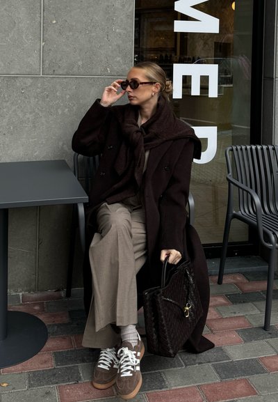 Abrigo marrón oscuro con una bufanda a juego, pantalones beige y zapatillas marrón claro. Sostenido un bolso de mano tejido oscuro, sentado en una mesa.