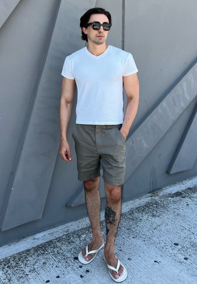 Camiseta blanca ajustada, pantalones cortos verdes oliva y chanclas blancas. El modelo está de pie contra una pared texturizada de color gris. Tatuaje visible en la pierna izquierda.
