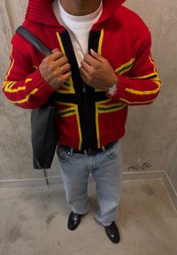 Άτομο που φοράει κόκκινο πλεκτό πουλόβερ με φερμουάρ και κίτρινες και πράσινες ρίγες, λευκό πουκάμισο, ανοιχτό μπλε τζιν, μαύρα παπούτσια, ρολόι χειρός και κρατά μαύρη τσάντα.