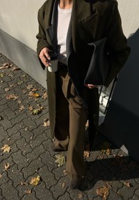 Ελιά πράσινο μακρύ παλτό, λευκή ριμπέ μπλούζα, καφέ παντελόνι με φαρδιά μπατζάκια, μαύρη υφασμάτινη τσάντα, κρατώντας ένα φλυτζάνι καφέ, αστικό σκηνικό με πεσμένα φύλλα.