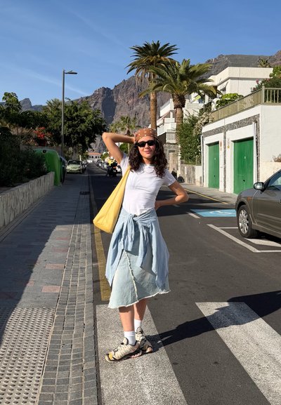 Mujer con gafas de sol y pañuelo en la cabeza posa en una calle iluminada por el sol con palmeras, montañas y coches aparcados al fondo.