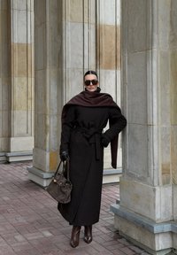 Ilgs melns mētelis ar brūnu šalli, ādas cimdi, saulesbrilles un brūna rokassoma. Stāv pret akmens kolonām uz ķieģeļu virsmas.