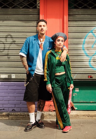 Dos personas están de pie frente a una pared cerrada. Una lleva una camisa azul, pantalones cortos negros y zapatos de cordones. La otra lleva un chándal verde.